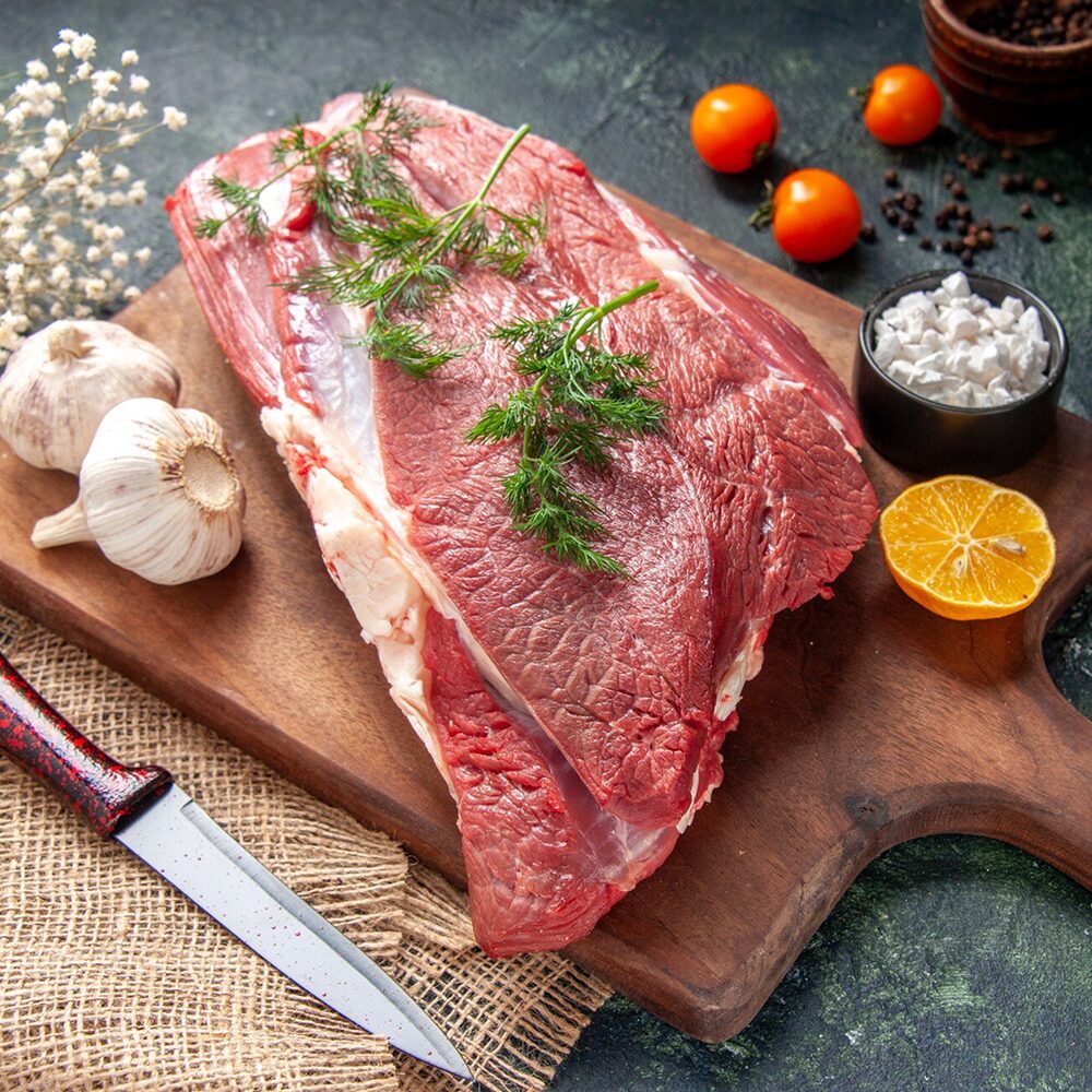 Front_view_of_fresh_raw_red_meats_green_garlics_lemon_salt_on_brown_wooden_cutting_board_knife_on_nude_color_towel_tomatoes_pepper_on_dark_color_background.jpg